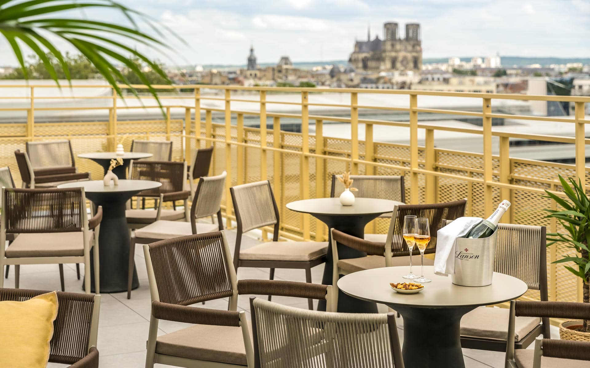 Rooftop terrace with outdoor seating, featuring round tables and woven armchairs distributed across the foreground and center, a champagne bucket with flutes on a table to the right, and a golden metal railing framing panoramic city views in the background; a cathedral silhouette rises on the horizon beneath a cloudy sky.