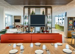 On-site dining area with light wood flooring, featuring a large communal table set with plates and cups in the foreground, bright red banquette seating at the center, and a buffet and beverage station visible in the background on the right; open shelving, contemporary furnishings, and abundant natural light create a welcoming restaurant atmosphere.