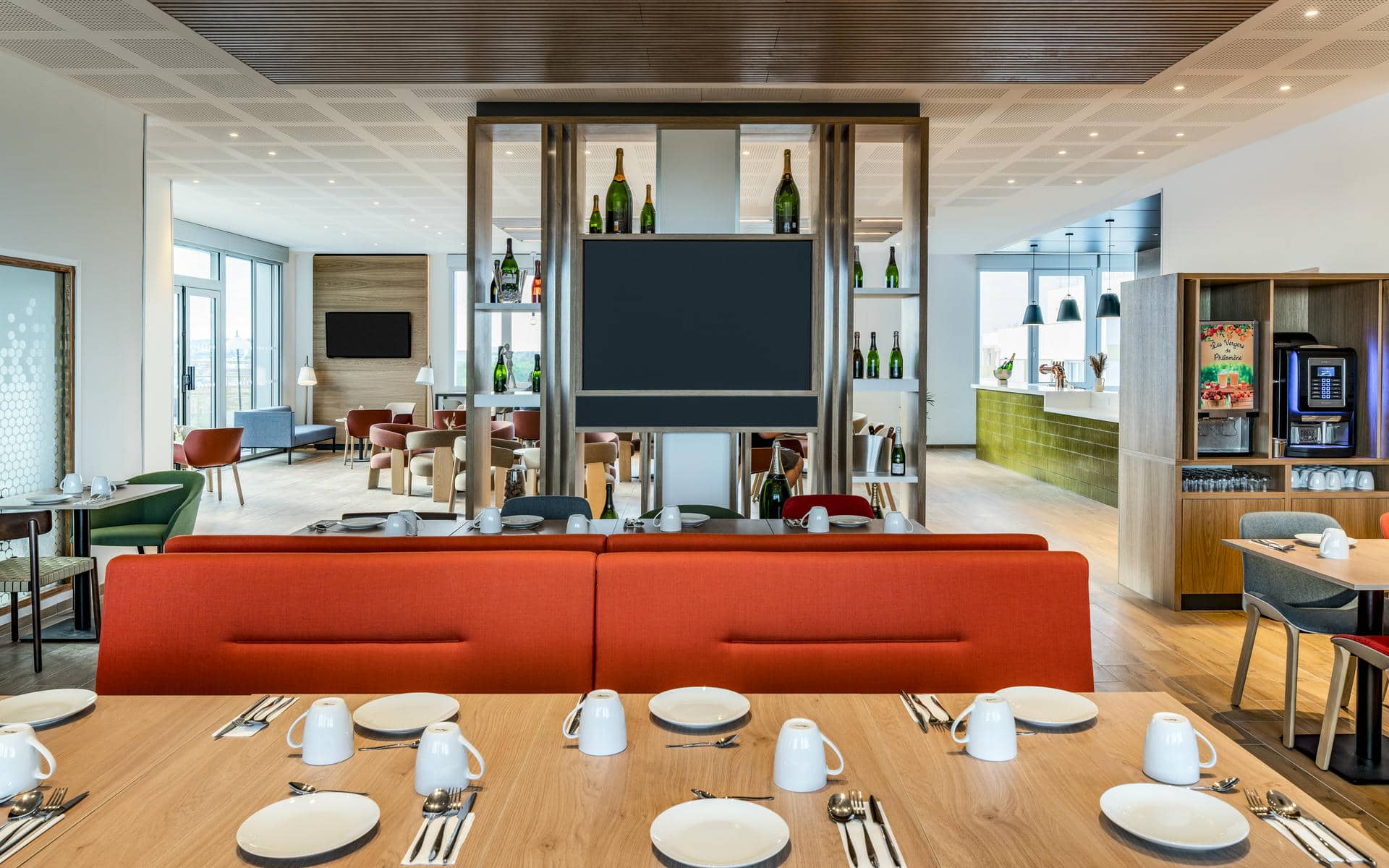 On-site dining area with light wood flooring, featuring a large communal table set with plates and cups in the foreground, bright red banquette seating at the center, and a buffet and beverage station visible in the background on the right; open shelving, contemporary furnishings, and abundant natural light create a welcoming restaurant atmosphere.