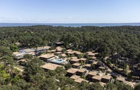 Aerial view of a hotel estate nestled within a vast pine forest, featuring clusters of lodge-style buildings with terracotta roofs and a central swimming pool across the foreground and center, while the sea stretches across the horizon in the background under a clear blue sky.
