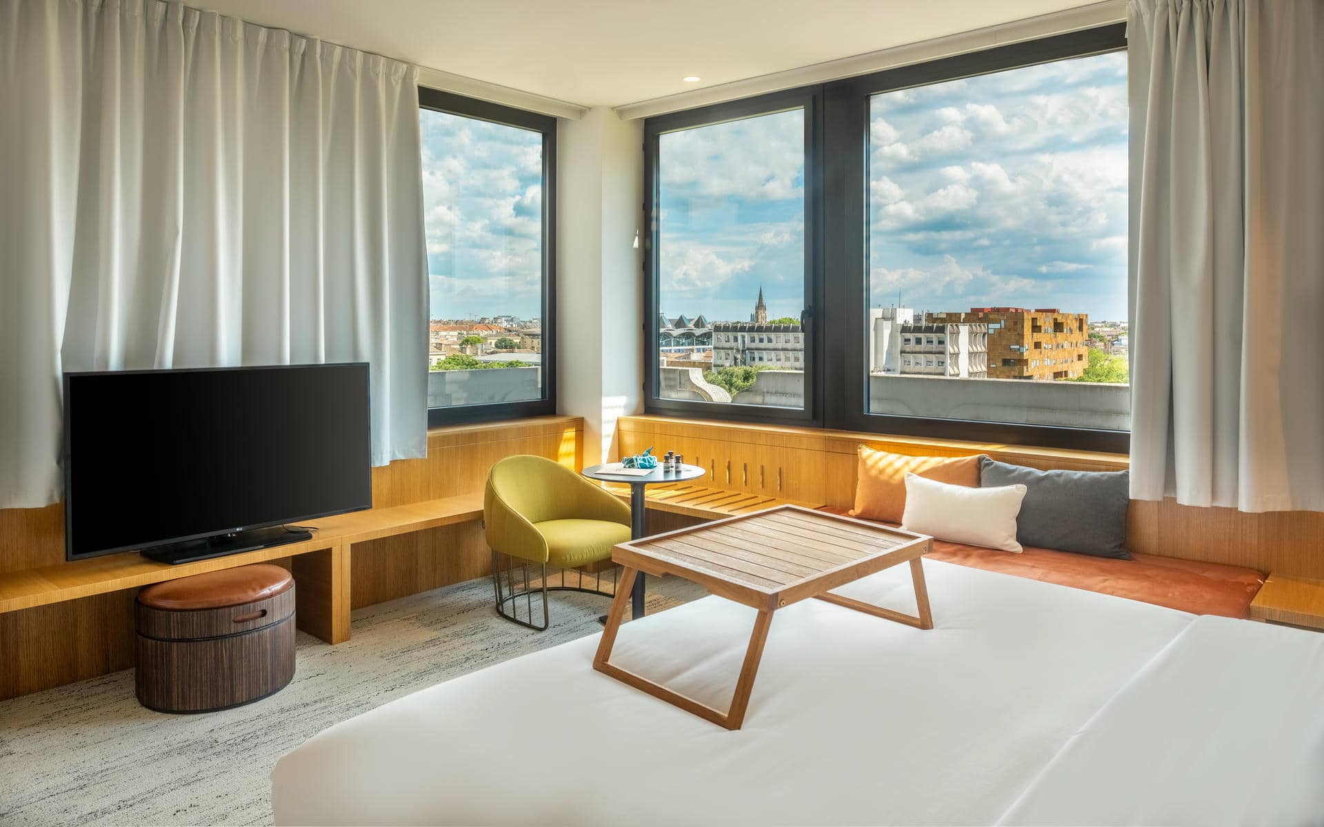 Bright hotel room with a bed in the foreground topped by a wooden tray, facing a corner seating area with a wooden window bench, a black round side table and a green armchair; large corner windows offer panoramic city views, while a television stands on the left.