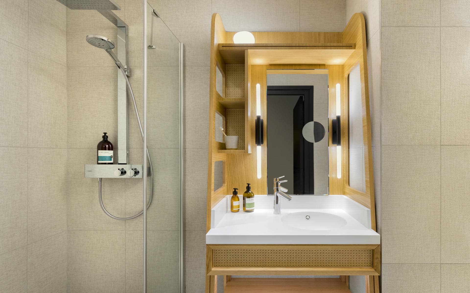 Hotel bathroom with light beige tiled walls, featuring on the left a glass-enclosed walk-in shower with a handheld showerhead and toiletries, and on the right a wooden vanity unit with open shelving, a white sink and a large illuminated mirror creating a bright, modern atmosphere.