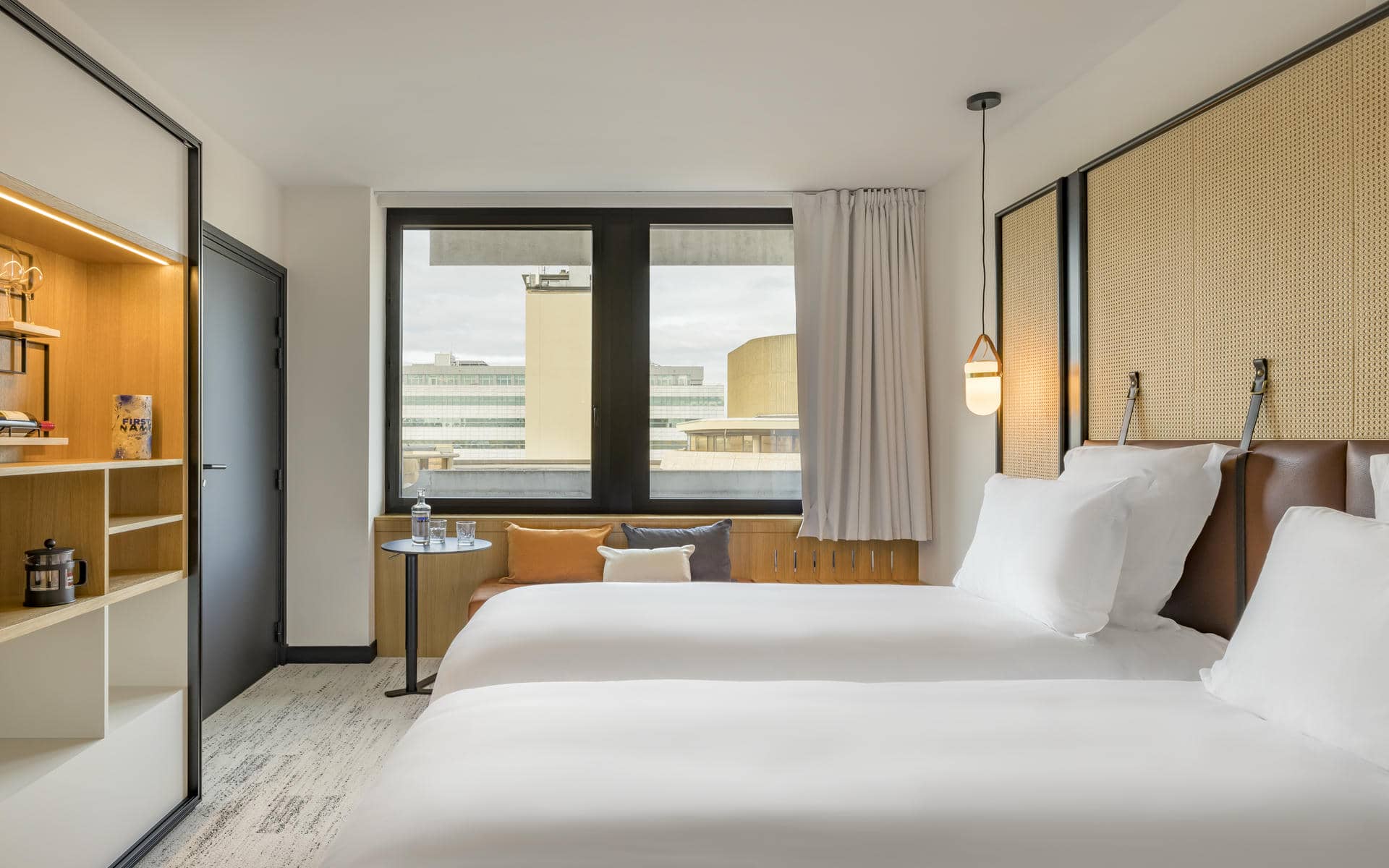 Hotel room with light-toned décor, featuring in the foreground two single beds with white bedding and woven-panel headboards, a black-framed window at the back overlooking urban buildings, and on the left a built-in shelving unit with warm lighting beside a small seating area with an armchair and side table.