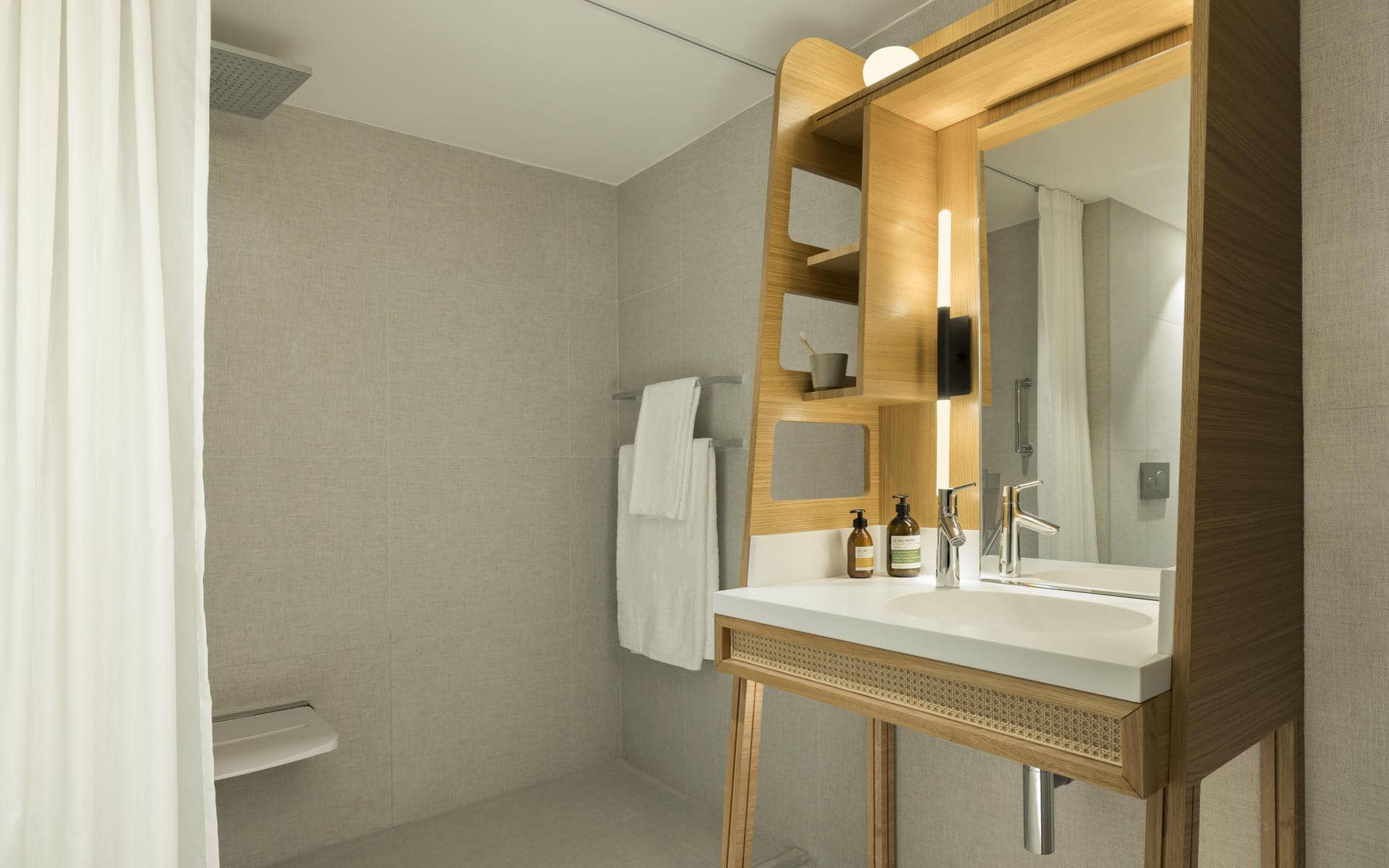 Hotel bathroom with a minimalist beige design, featuring on the right a wooden vanity unit with an illuminated mirror and integrated washbasin, while on the left a walk-in shower with a ceiling-mounted showerhead extends along a plain tiled wall with white towels.