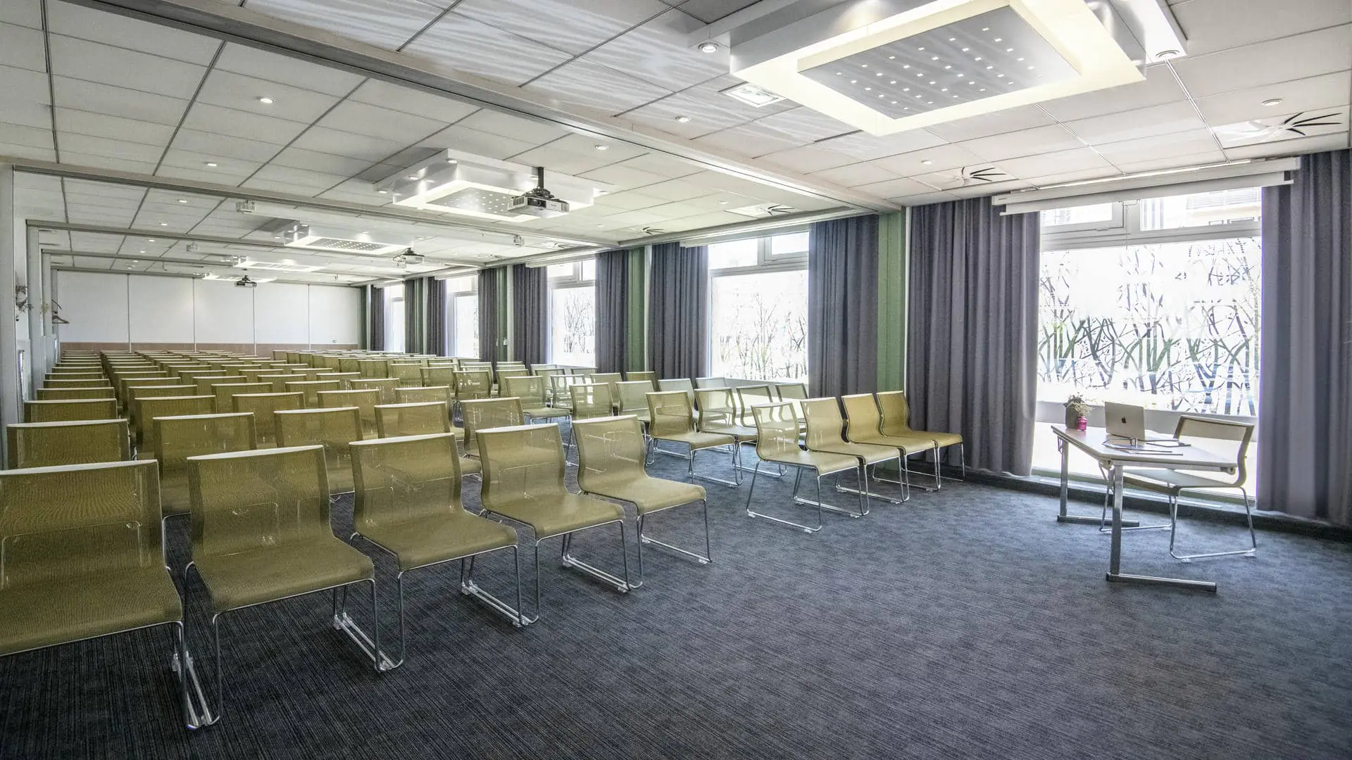 White and olive-green chairs arranged in front of a desk, with a projection screen facing them and five windows on the right side