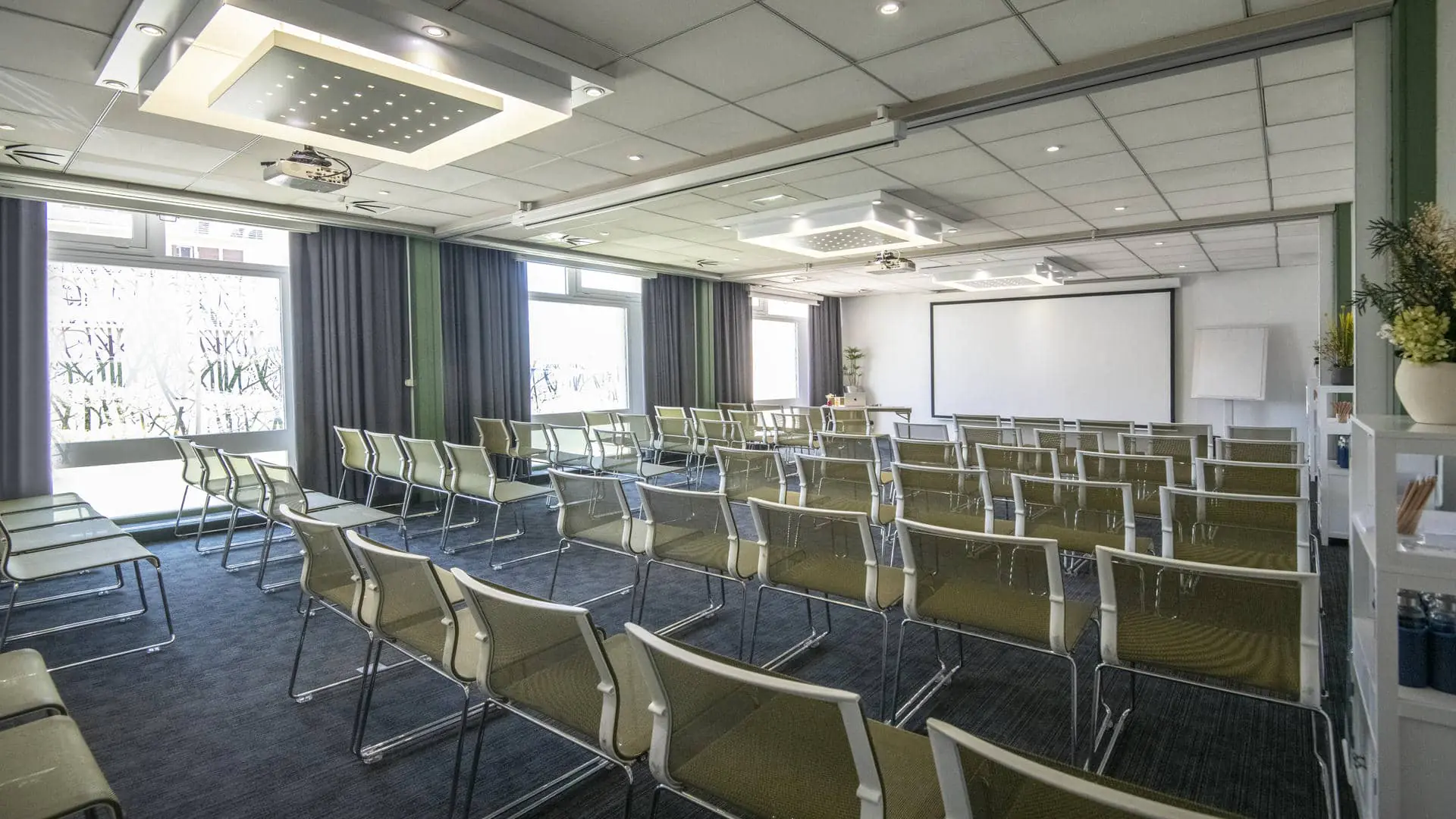 White and olive-green chairs arranged in front of a desk, with a projection screen facing them and three windows on the right side