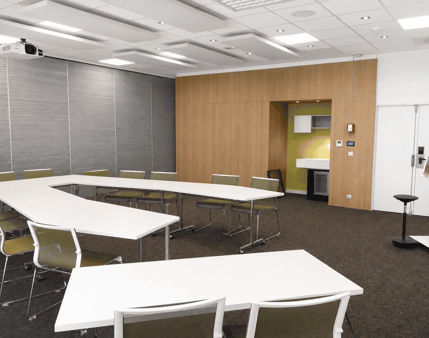 Seminar room in Saint-Quentin-en-Yvelines with white U-shaped tables, white and green chairs, grey carpet, one grey wall section and another wall section clad in wood.