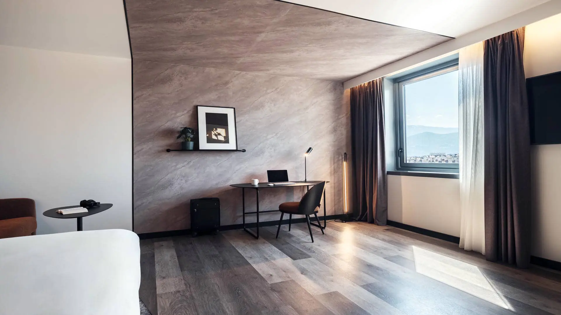 View from the double bed towards the window, with a mountain view. The sleeping area is separated from the work area by a black line, with different floor coverings and contrasting wall finishes on each side.