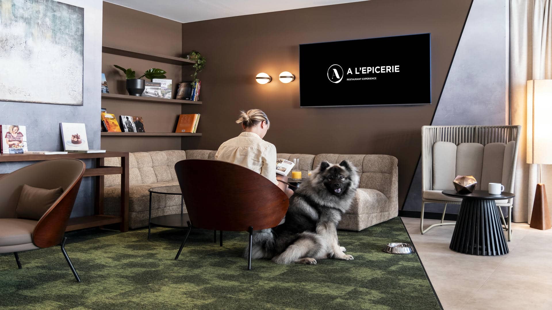 Woman seen from behind reading a book in a cosy lounge at a seminar hotel in Grenoble, while a dog looks at the camera, with a large TV screen mounted on the wall.