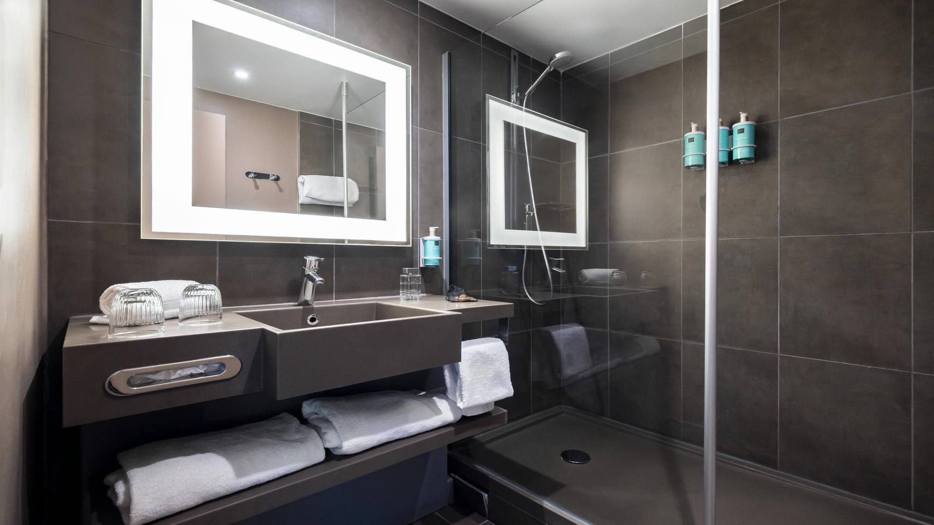 Brown tiled shower room with a large vanity unit, two washbasins, and backlit mirrors.