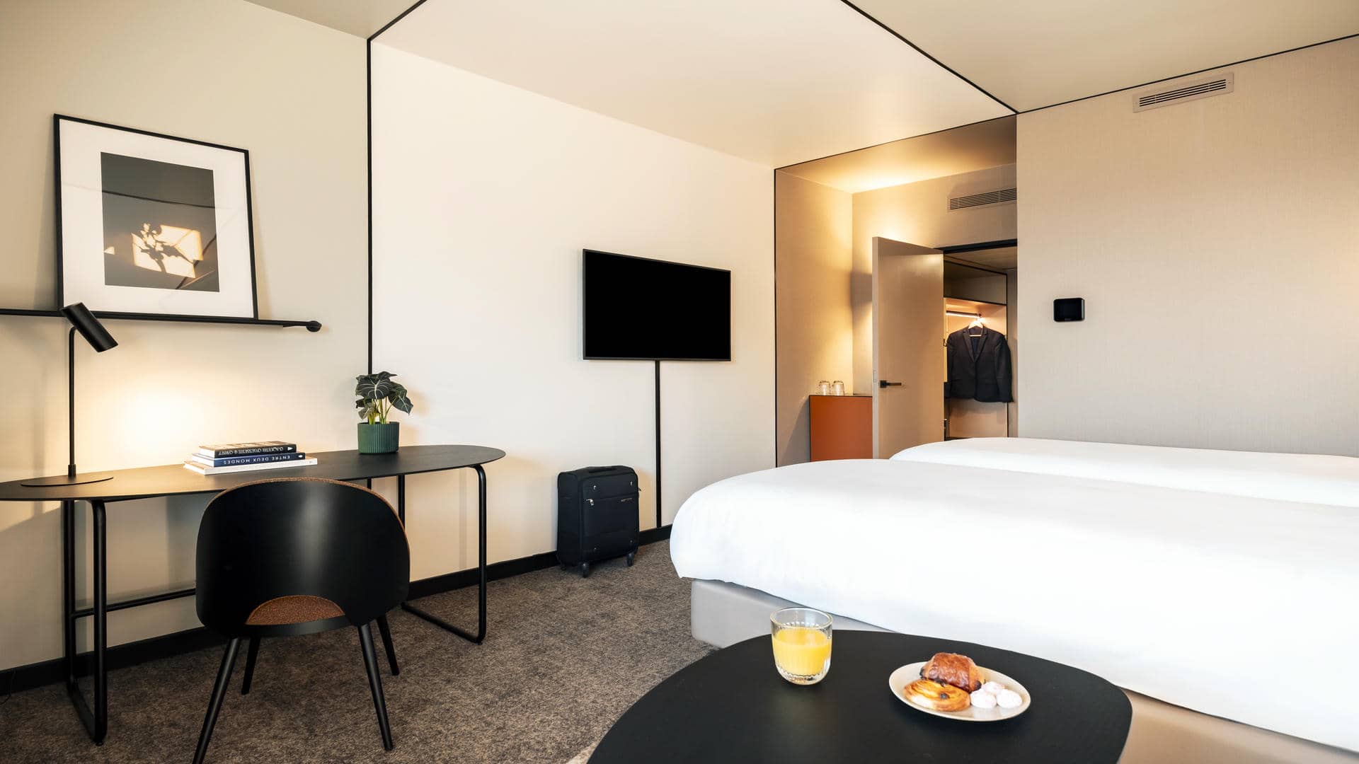 Double bed in twin configuration, with a table and chair beside it, featuring fruit juice and pastries. A dressing area is visible from the shooting angle, with a wall-mounted TV screen and a suitcase underneath.