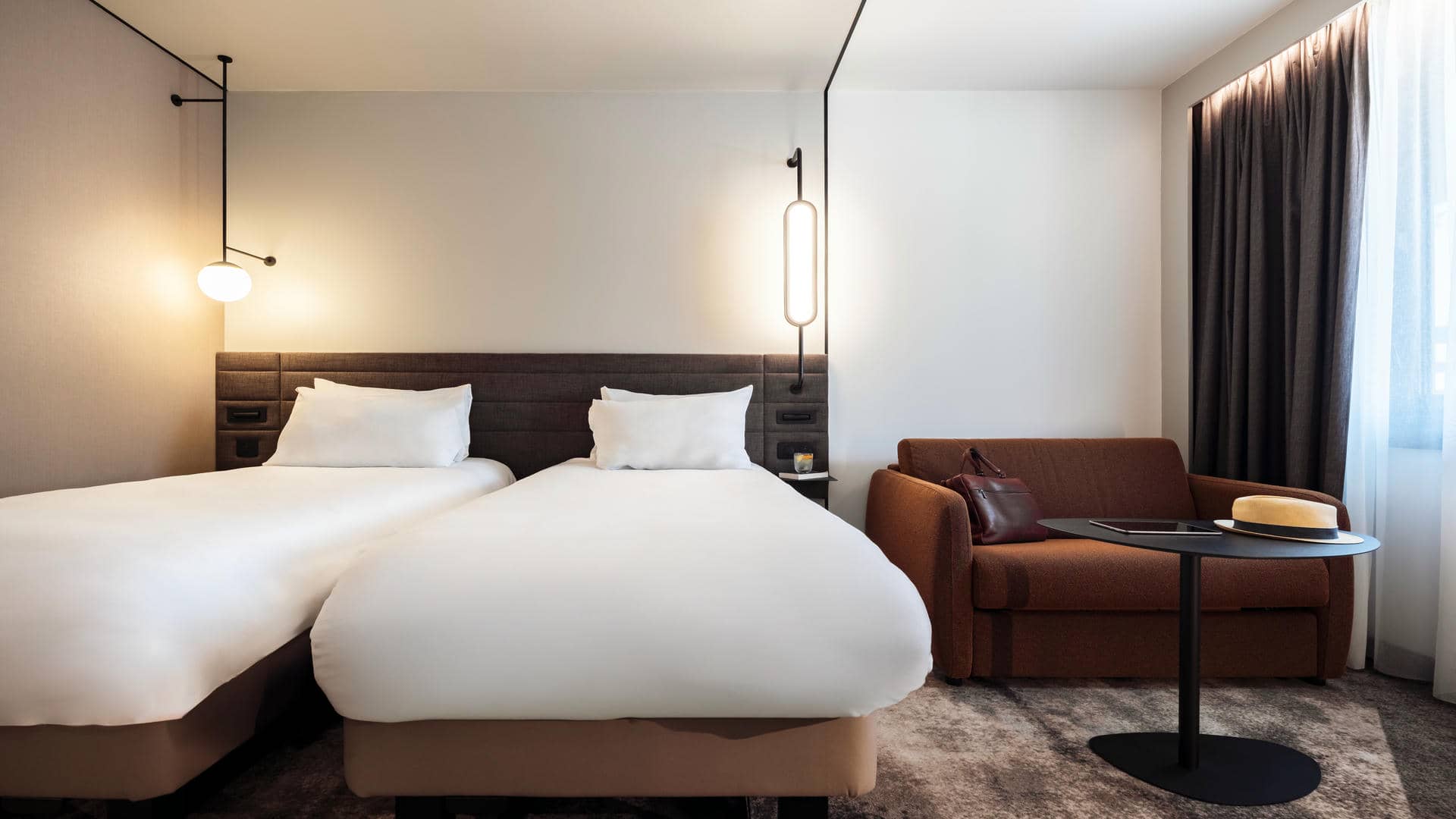 Twin beds with a single sofa bed beside them, a work bag placed on top, and a round table with a hat and tablet.