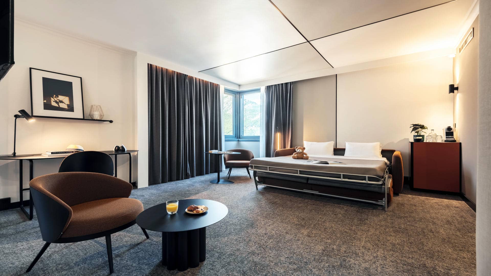 Lounge area of the Novotel Grenoble suite, featuring an orange double sofa bed shown open to accommodate two additional guests alongside the double bedroom, a work table decorated with a design photograph and decorative objects, and a corner window with thick double curtains.