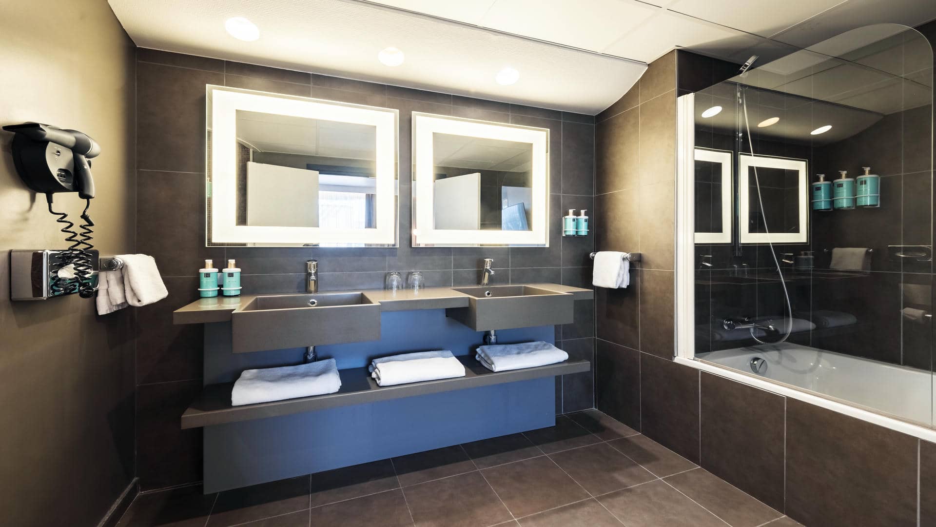 Brown tiled bathroom with a large vanity unit, two washbasins, and backlit mirrors.