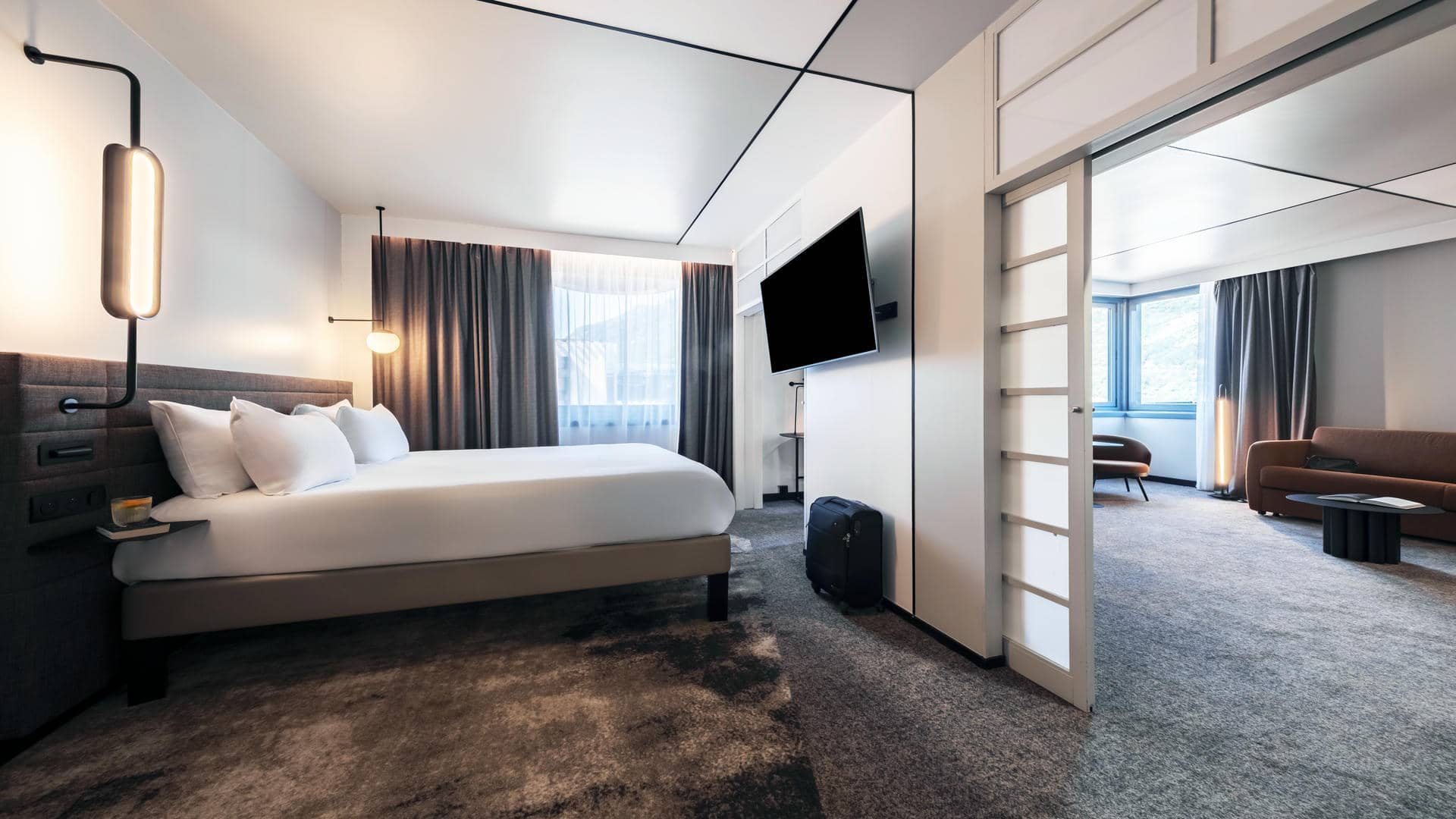 Design suite at the Novotel Grenoble, featuring dual carpeting to distinguish the sleeping area from the lounge area, sliding doors, and a mountain view in brown and orange tones.