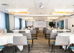 en::Reception venue set up in banquet style, with round tables and floral centrepieces, facing a screen displaying Novotel Grenoble Centre. fr::Lieu de réception présenté en banquet, avec tables rondes aux centres de table fleuris, face à un écran affichat Novotel Grenoble Centre.