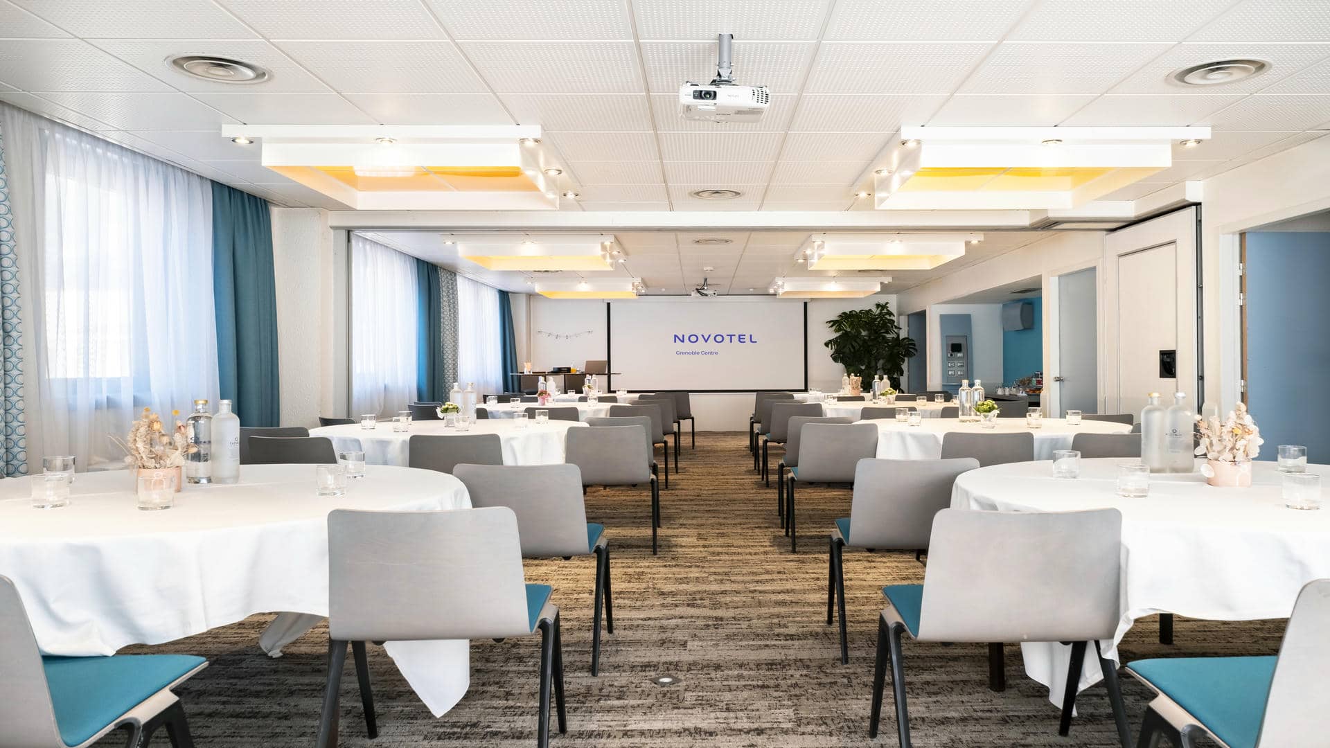 en::Reception venue set up in banquet style, with round tables and floral centrepieces, facing a screen displaying Novotel Grenoble Centre. fr::Lieu de réception présenté en banquet, avec tables rondes aux centres de table fleuris, face à un écran affichat Novotel Grenoble Centre.