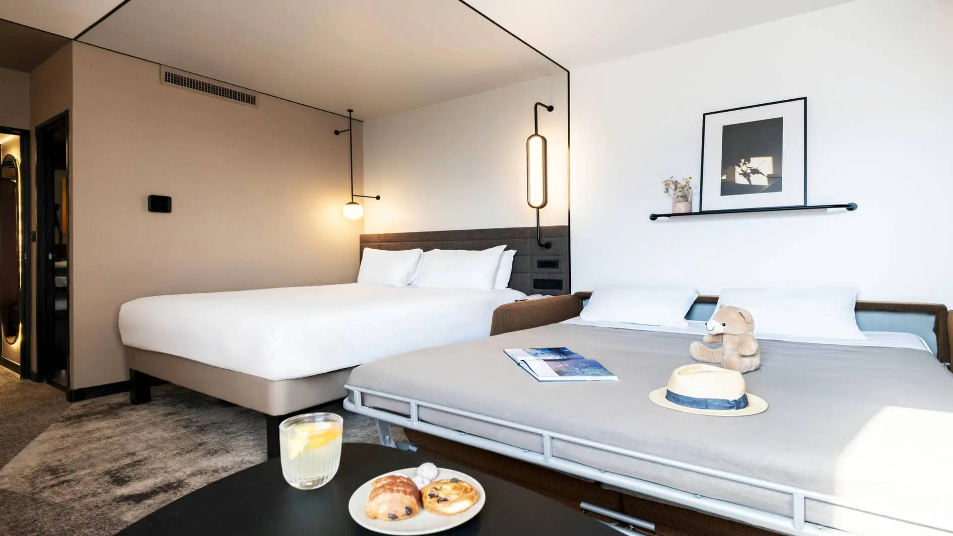Black design lights switched on above the double bed, next to an open sofa bed with a soft toy and magazine placed on it. On the other side of the room, a mirror and the bathroom door can be seen.
