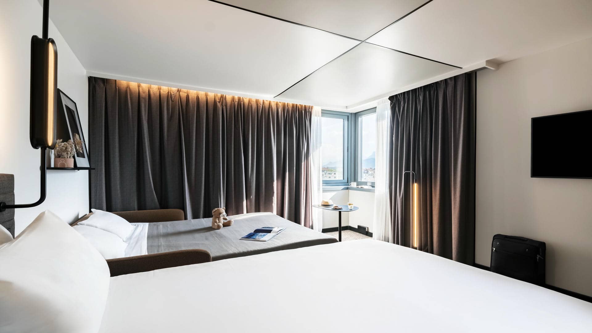 Brown curtains drawn along a corner window, with a black design light switched on above the double bed, next to an open sofa bed with a soft toy and magazine placed on it.
