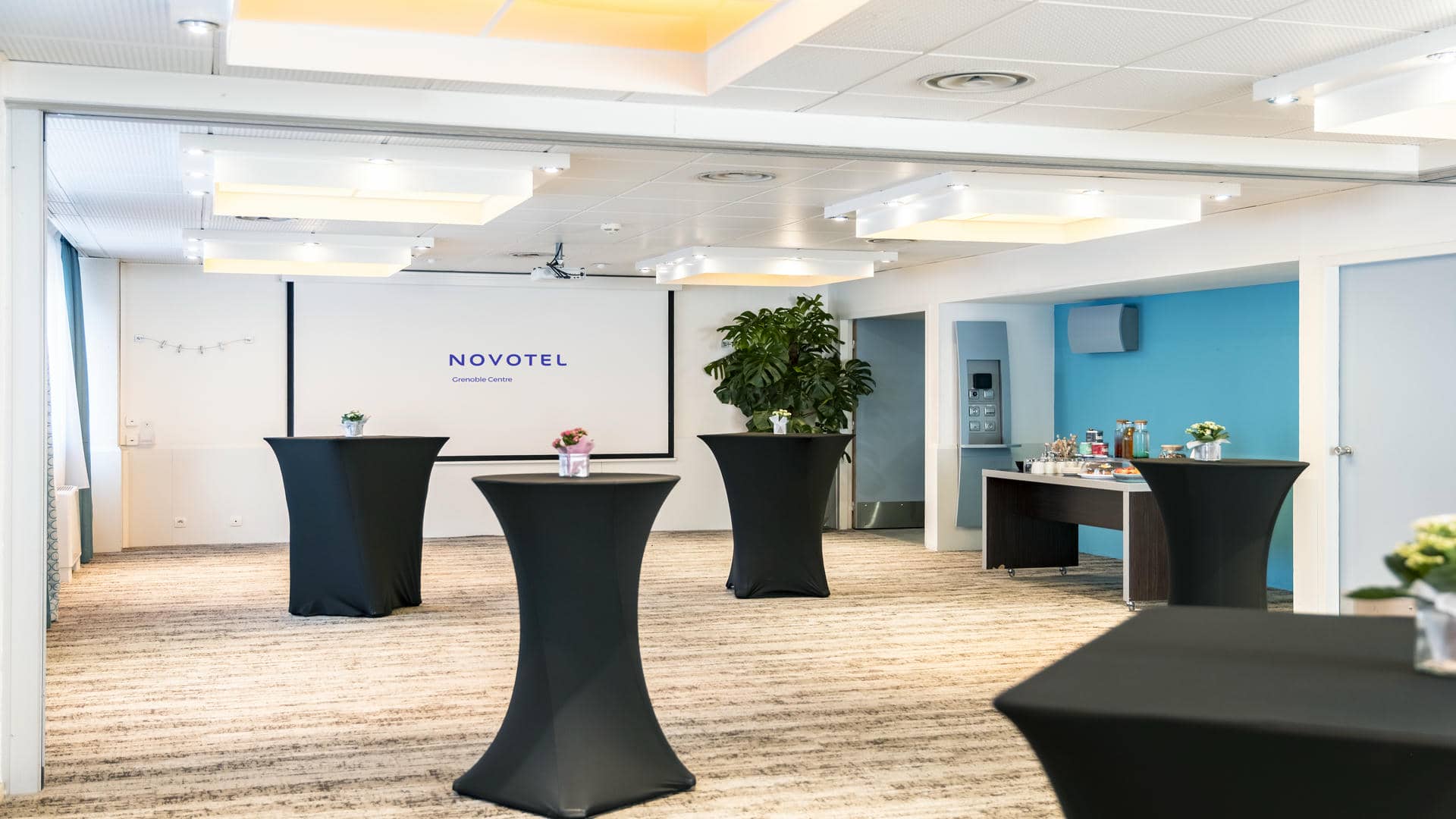 en::Bright reception room with a screen lowered against a white wall displaying the Novotel Grenoble Centre logo, set up with black-covered cocktail tables and floral centrepieces. fr::Salle de réception lumineuse avec écran descendu sur le mur blanc affichant le logo du Novotel Grenoble Centre, présentée avec des mange-debout habillés de noir et des centres de table fleuris.
