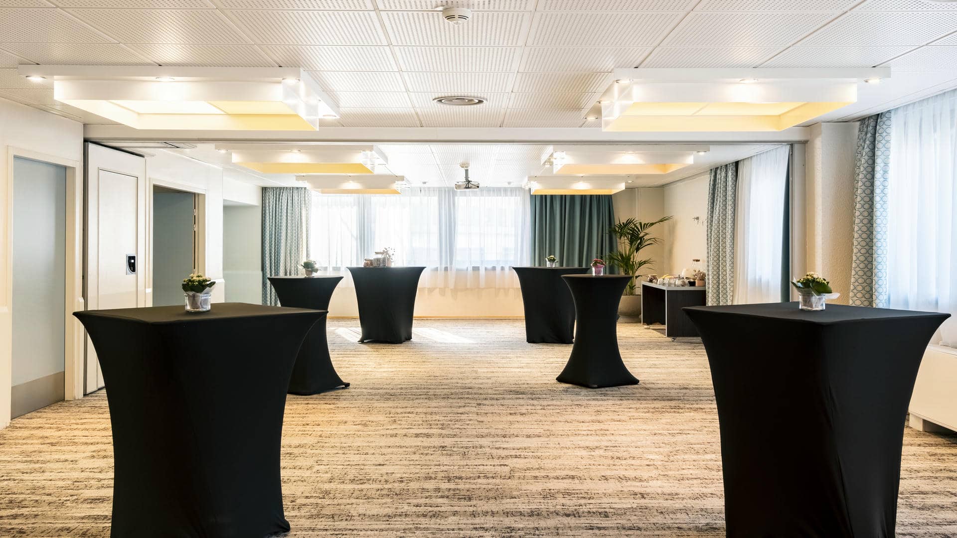 en::Bright reception room with green curtains and elegant grey carpet, set up with black-covered cocktail tables and floral centrepieces. fr::Salle de réception lumineuse aux rideaux verts et à la moquette grise élégante, présentée avec des mange-debout habillés de noir et des centres de table fleuris.