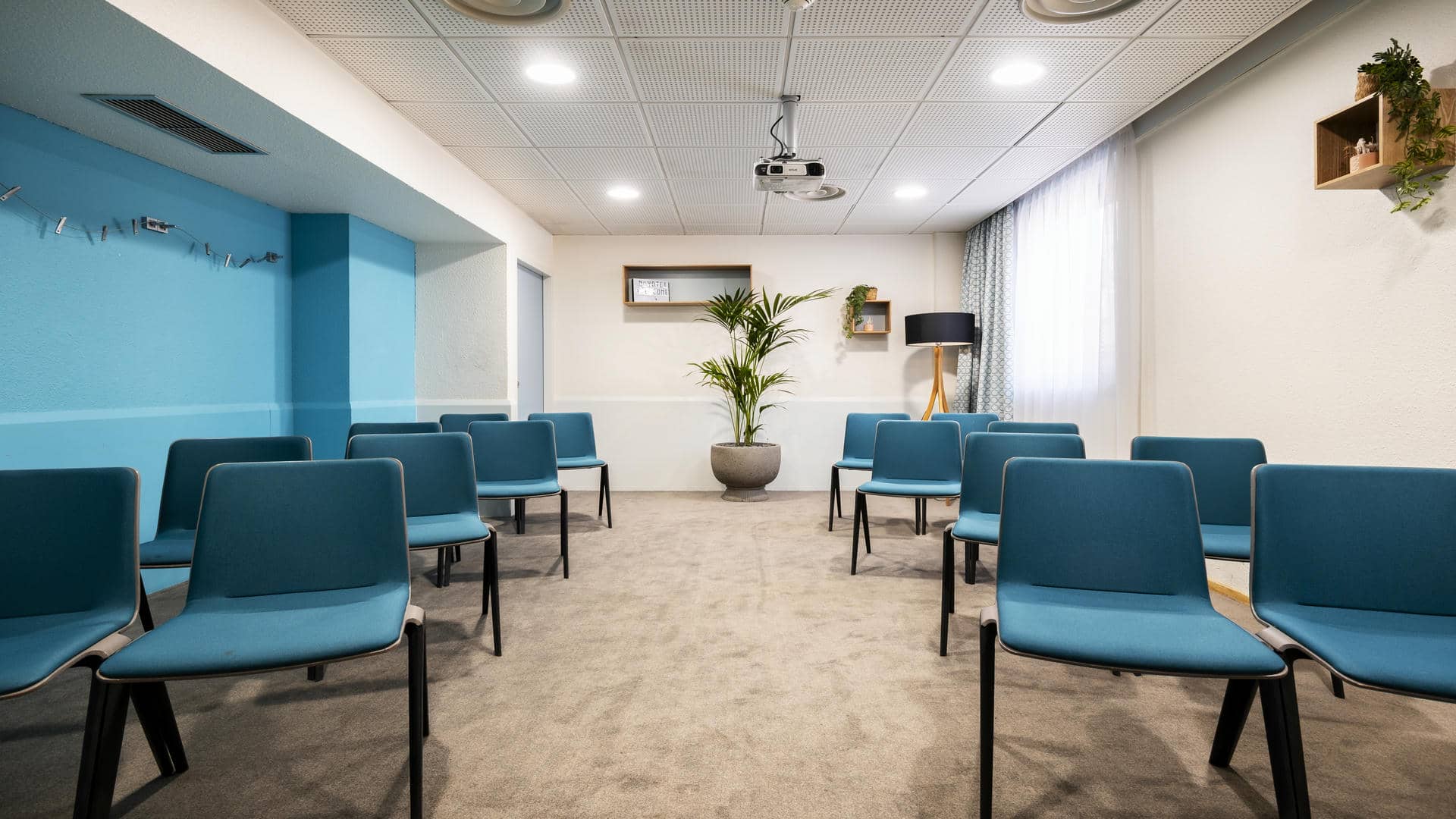 Air-conditioned training room in Grenoble, equipped with a projection screen, featuring blue and grey chairs, a wall in matching shades of blue, a large window, and soft-toned décor. fr: Salle de formation à louer au Novotel Grenoble, équipée d’un rétroprojecteur, avec des chaises bleues et grises, un mur dans des tons de bleu assortis, une grande fenêtre et une décoration aux tons doux.
