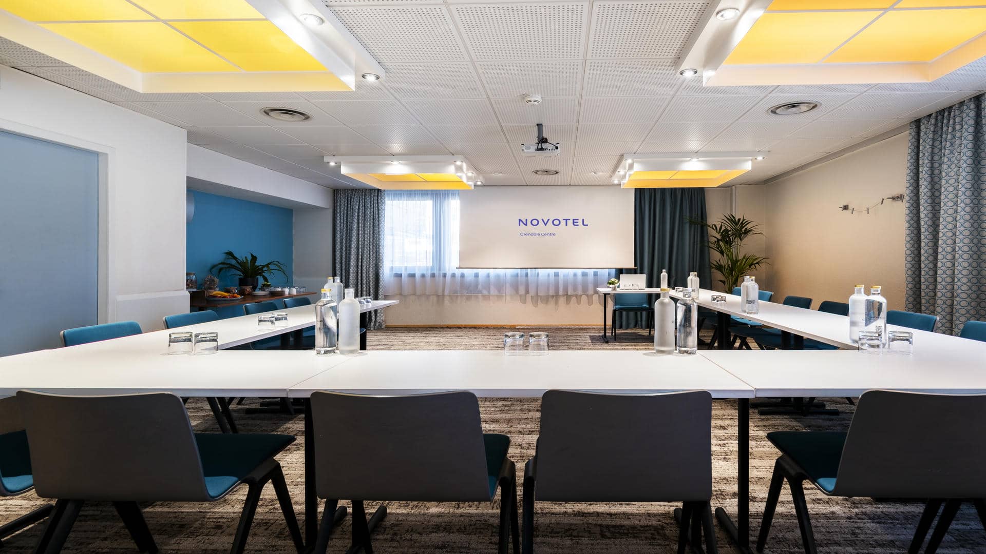Meeting room in Grenoble available for hire, set up in a U-shape facing a screen lowered in front of a large window, with yellow ceiling lights and contemporary décor.
