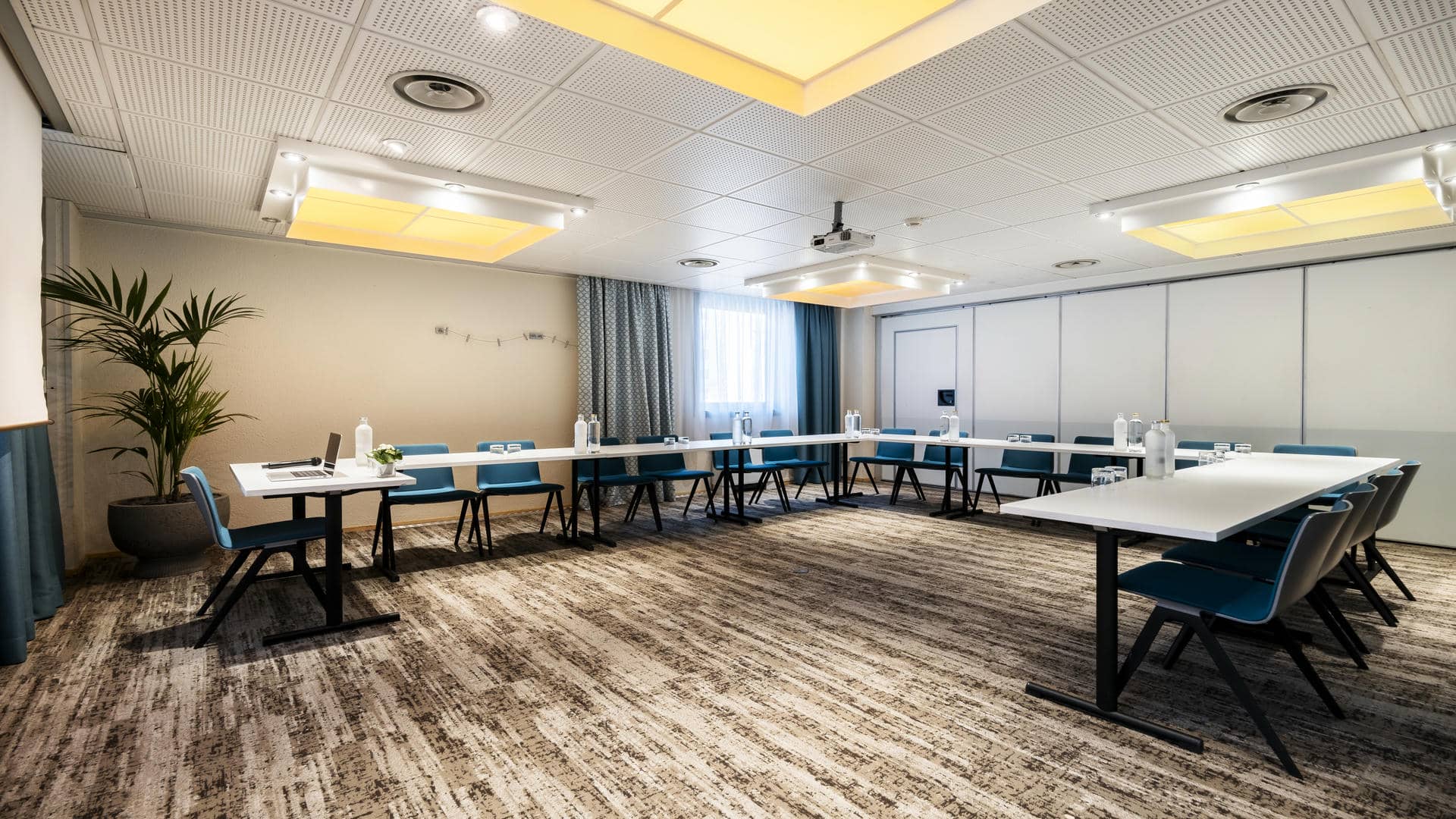 en::Training room with a desk equipped with a microphone, a lowered screen behind it and U-shaped meeting tables facing it. fr::Salle de formation avec bureau équipé d’un micro, écran descendu en arrière-plan et tables de réunion disposées en U face au bureau.