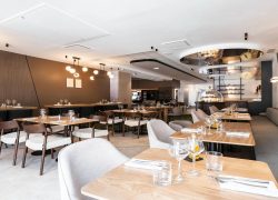 The seminar restaurant is photographed from the street-side entrance towards the back of the room, with square wooden tables and higher tables, along with sofa seating and fabric chairs.