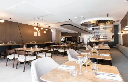 The seminar restaurant is photographed from the street-side entrance towards the back of the room, with square wooden tables and higher tables, along with sofa seating and fabric chairs.