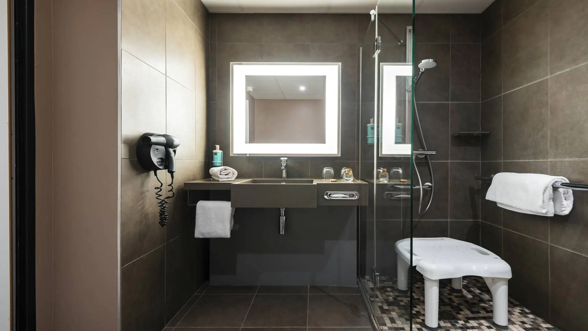 Accessible brown tiled shower room for guests with reduced mobility, featuring a large vanity unit, one washbasin, and a hairdryer.