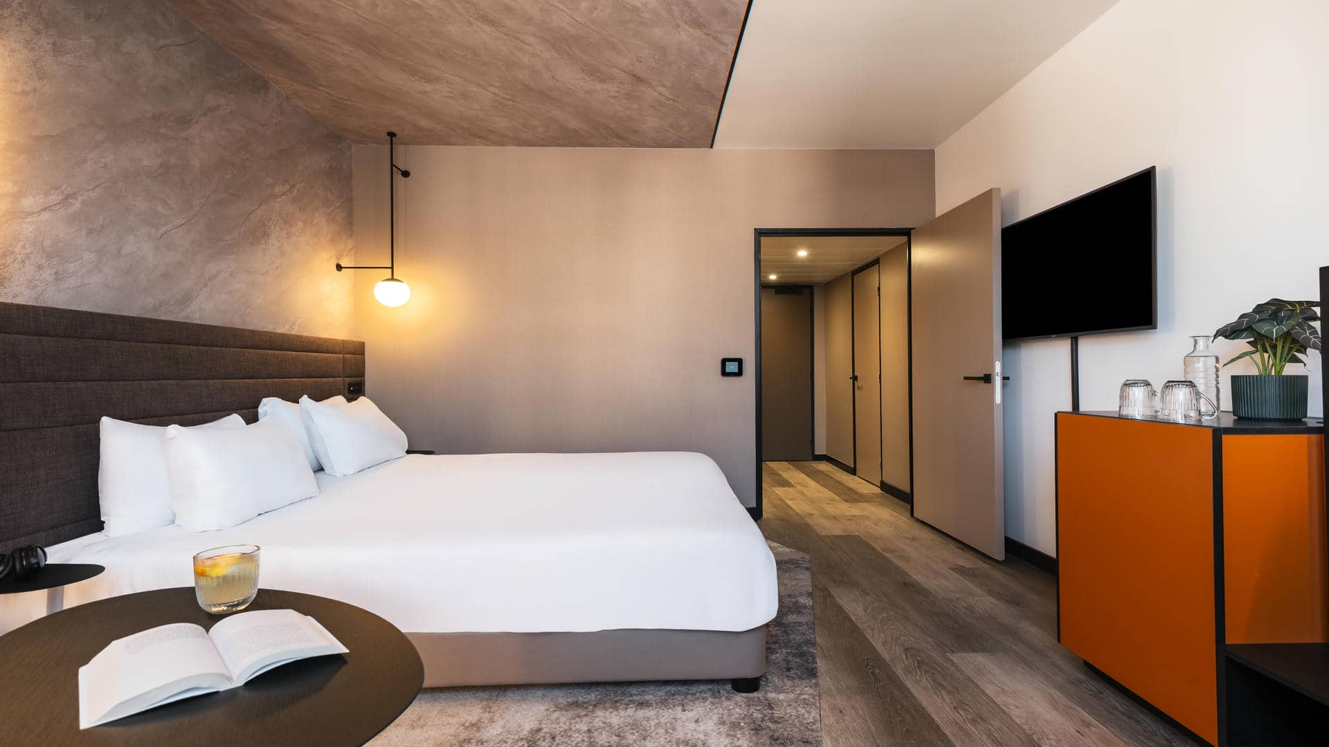 Double bed with a brown headboard, adjoining wall and ceiling covered in pink, green, and brown marble-effect wallpaper, with an open door leading towards the bathroom and entrance door.