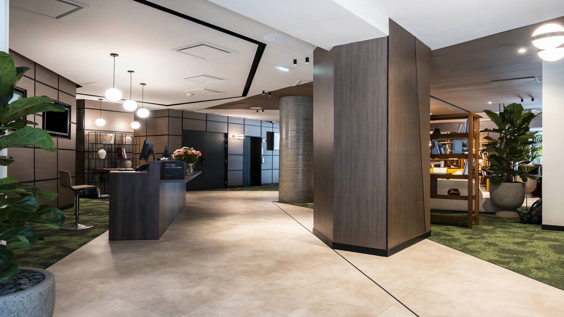 en::Reception area of the Novotel Grenoble Centre, with designer brown desks on the left, access to the lifts in the background and a large lobby opening onto the bar behind tall columns on the right. fr::Réception du Novotel Grenoble Centre avec desks marron design à gauche, accès aux ascenseurs au fond et grand lobby ouvert sur le bar derrière de hautes colonnes à droite.