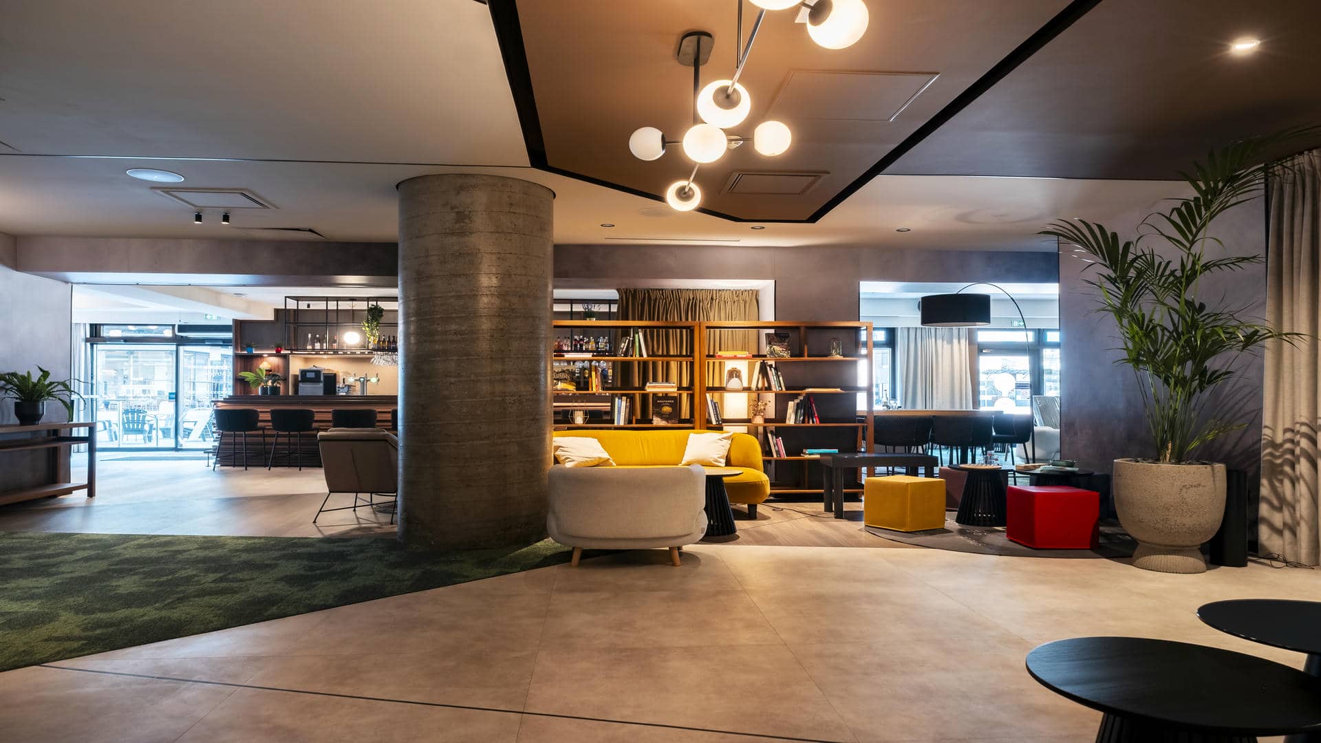 Large wooden counter with high chairs spanning much of the room’s width, with a lounge and library area in the foreground, and access to the outdoor terrace behind the bar in the background.