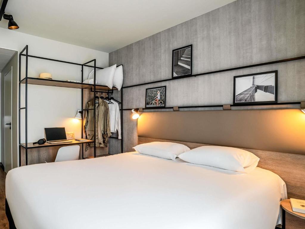 Hotel double bed room with a grey concrete-effect wall, featuring a beige upholstered headboard with integrated reading sconces and three black-framed black and white photographs above, a white bed in the foreground, and an open metal shelving unit with a clothes rail, desk and laptop on the left, for a seminar hotel stay.