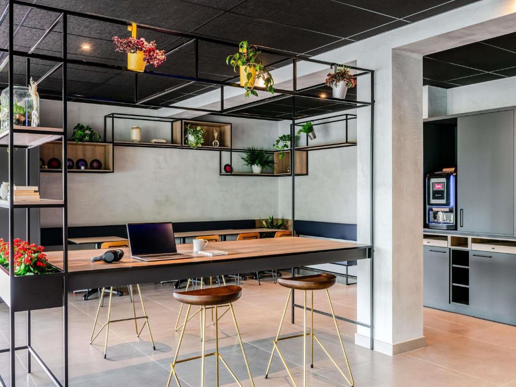 Hotel common workspace with a black ceiling and tiled floor, featuring a long wooden high table with gold-legged wooden bar stools, a laptop and headphones on the surface, framed by a black metal shelving unit with plants and decorative objects, red flowering plants on the left, and a grey kitchenette area with a coffee machine on the right.