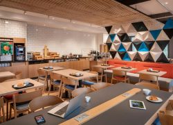 Vue générale de la salle de petit déjeuner d'hôtel avec plafond à lames de bois, comprenant des tables et chaises en bois clair avec viennoiseries et tasses au premier plan, une banquette rouge contre un mur de panneaux géométriques 3D bleu, noir et blanc à droite, et un comptoir buffet avec carrelage métro blanc et meubles en bois à gauche.