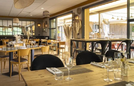 General view of a restaurant dining room with wooden flooring, featuring tables set with glassware and light wood chairs across the foreground and center, black upholstered chairs on the right, and large openings onto a covered terrace in the background; woven light fixtures, decorative dried grasses, and warm-toned furnishings create a contemporary atmosphere.