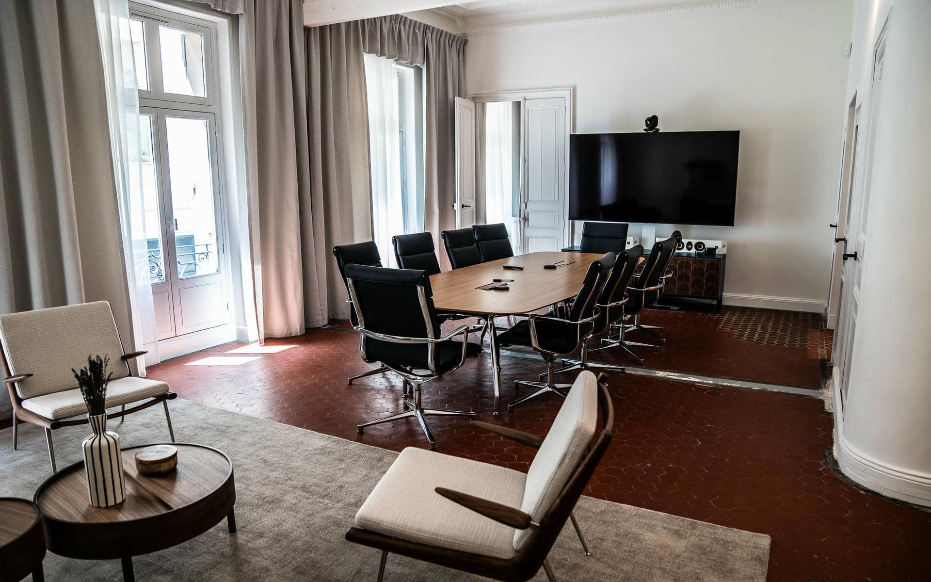 Salle de location pour réunion dans un cadre haussmannien avec un sol en tomettes hexagonales terracotta bordeaux, une table de conférence ovale en noyer entourée de chaises en cuir noir et chrome face à un grand écran plat sur le mur de droite, deux fauteuils beige avec une table basse ronde en bois sur un tapis gris au premier plan, de hautes fenêtres françaises avec rideaux en lin gris à gauche, et des murs blancs moulurés.