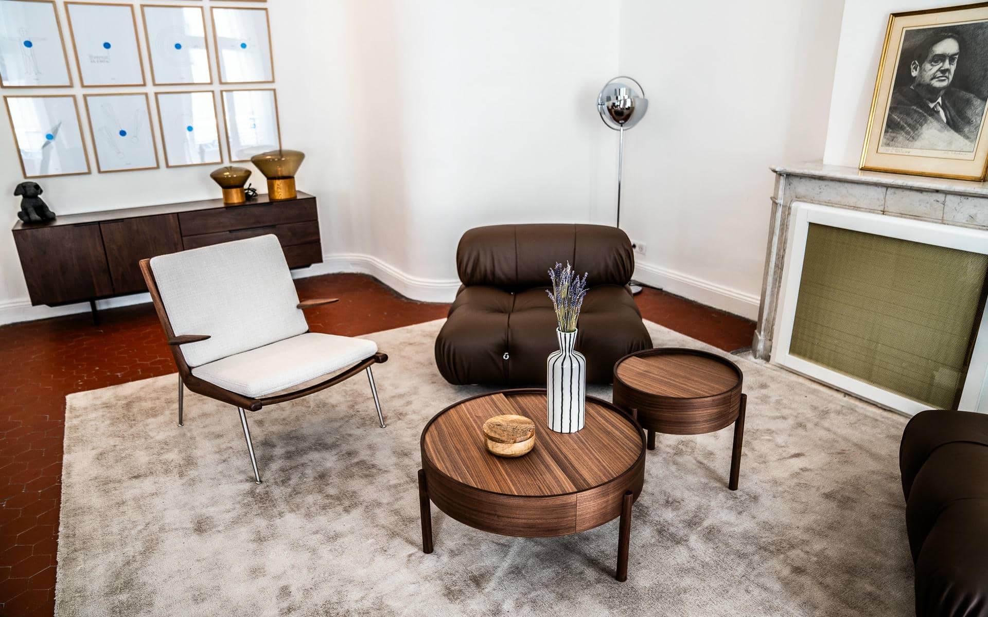 Informal meeting lounge with a terracotta floor and beige carpet, featuring a light grey fabric armchair with chrome legs on the left, a dark brown leather lounge chair in the centre, two round walnut coffee tables with a striped vase of lavender, a dark wood sideboard with decorative objects and a grid of framed prints on the back wall, a chrome globe floor lamp, and a marble fireplace surround on the right.