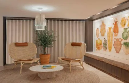 Hotel lobby lounge in a wide view, featuring at the center two woven rattan lounge chairs facing a round stone coffee table on a textured rug, with a potted tree in the background; on the right, a long built-in bench runs beneath a decorative wall mural of colorful ceramic vessels, while full-height curtains soften the space.