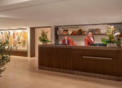 Hotel reception area with light hexagonal floor tiles, featuring a large dark wood reception desk occupying the foreground and center, two staff members behind the counter on the right, and a decorative ceramic mural with built-in seating along the left wall; open shelving, floral arrangements, and warm lighting create an elegant and welcoming atmosphere.