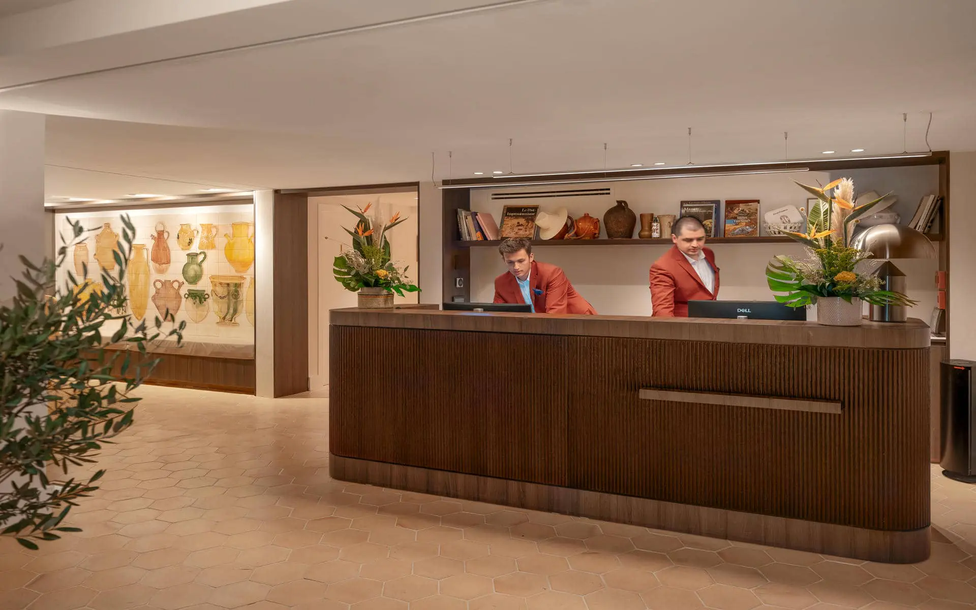 Hotel reception area with light hexagonal floor tiles, featuring a large dark wood reception desk occupying the foreground and center, two staff members behind the counter on the right, and a decorative ceramic mural with built-in seating along the left wall; open shelving, floral arrangements, and warm lighting create an elegant and welcoming atmosphere.
