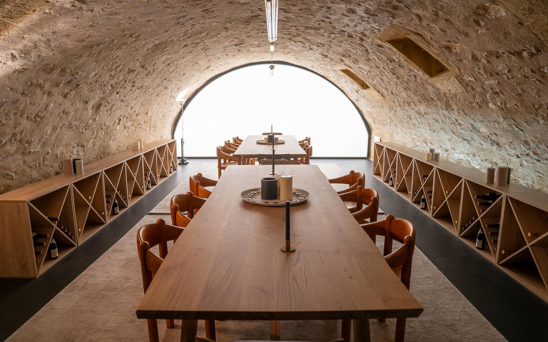 Unusual meeting room rental space in a vaulted stone cellar with two long oak tables and cognac-coloured wooden armchairs on a beige rug, wine bottle storage racks in light wood lining both sides, candles and a centrepiece on each table, a dark concrete floor, and a large illuminated arched opening at the far end.