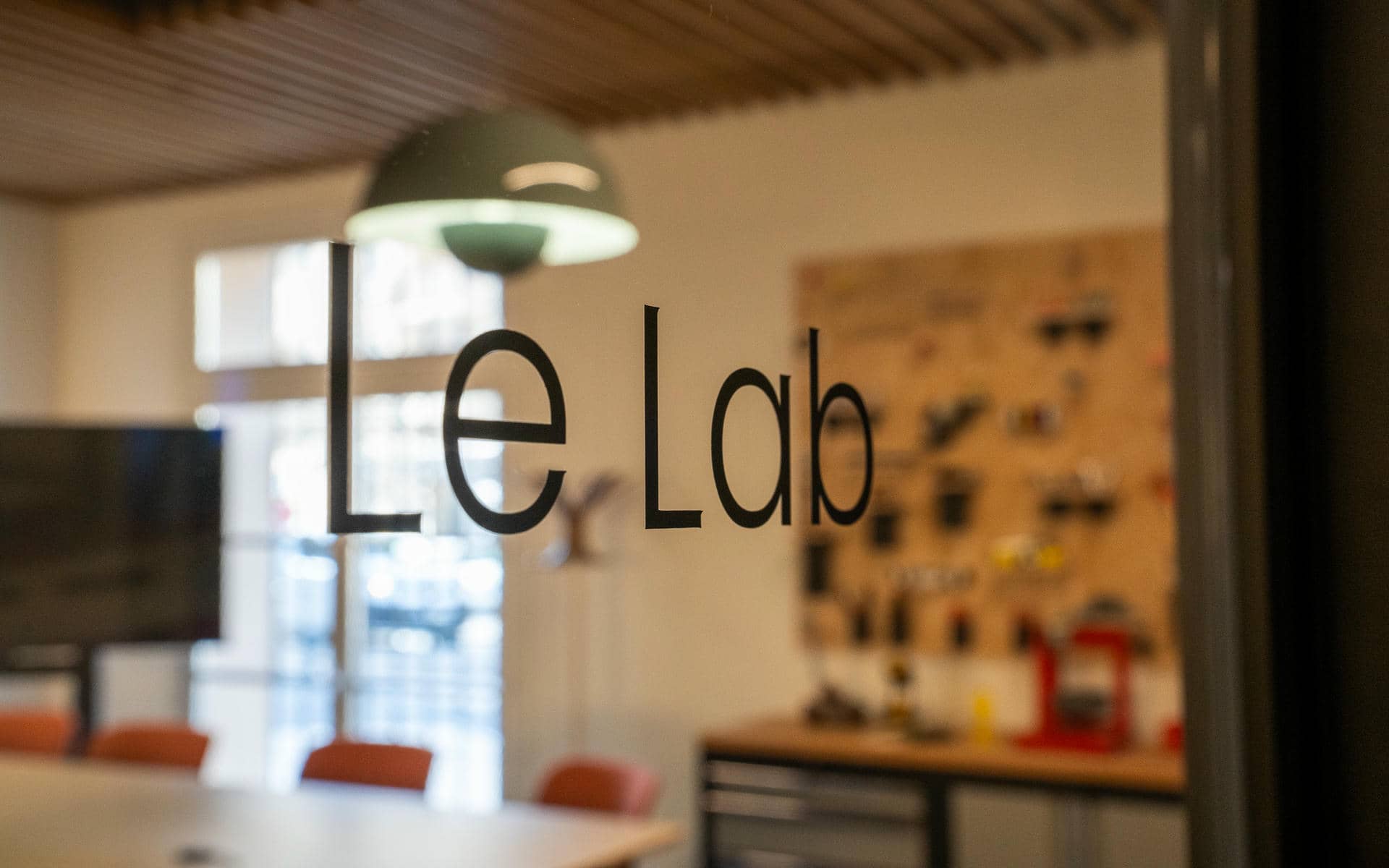 Close-up of a meeting room glass door with "Le Lab" in black lettering, revealing through the glass a wood-slatted ceiling, a green pendant lamp, orange chairs, a workbench with a red kitchen mixer and a pegboard wall with various tools and accessories in the background.