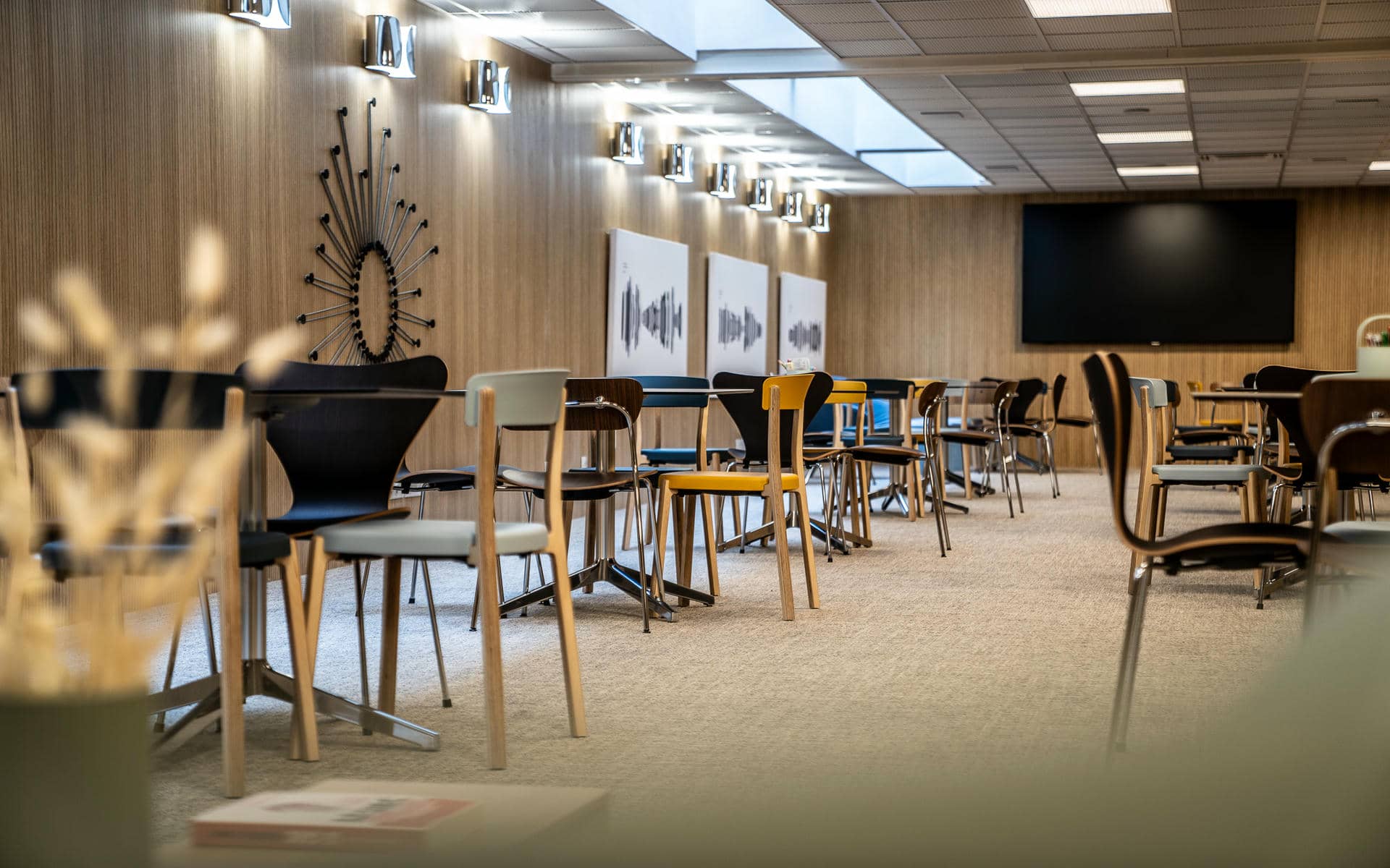 Training room with rows of dark rectangular tables and mixed design chairs in black, yellow and wood tones on a beige carpet, wood-panelled walls with a sunburst wall clock, a row of chrome wall sconces, three abstract art panels, and a large black flat screen at the far end, with a blurred dried flower arrangement in the foreground on the left.