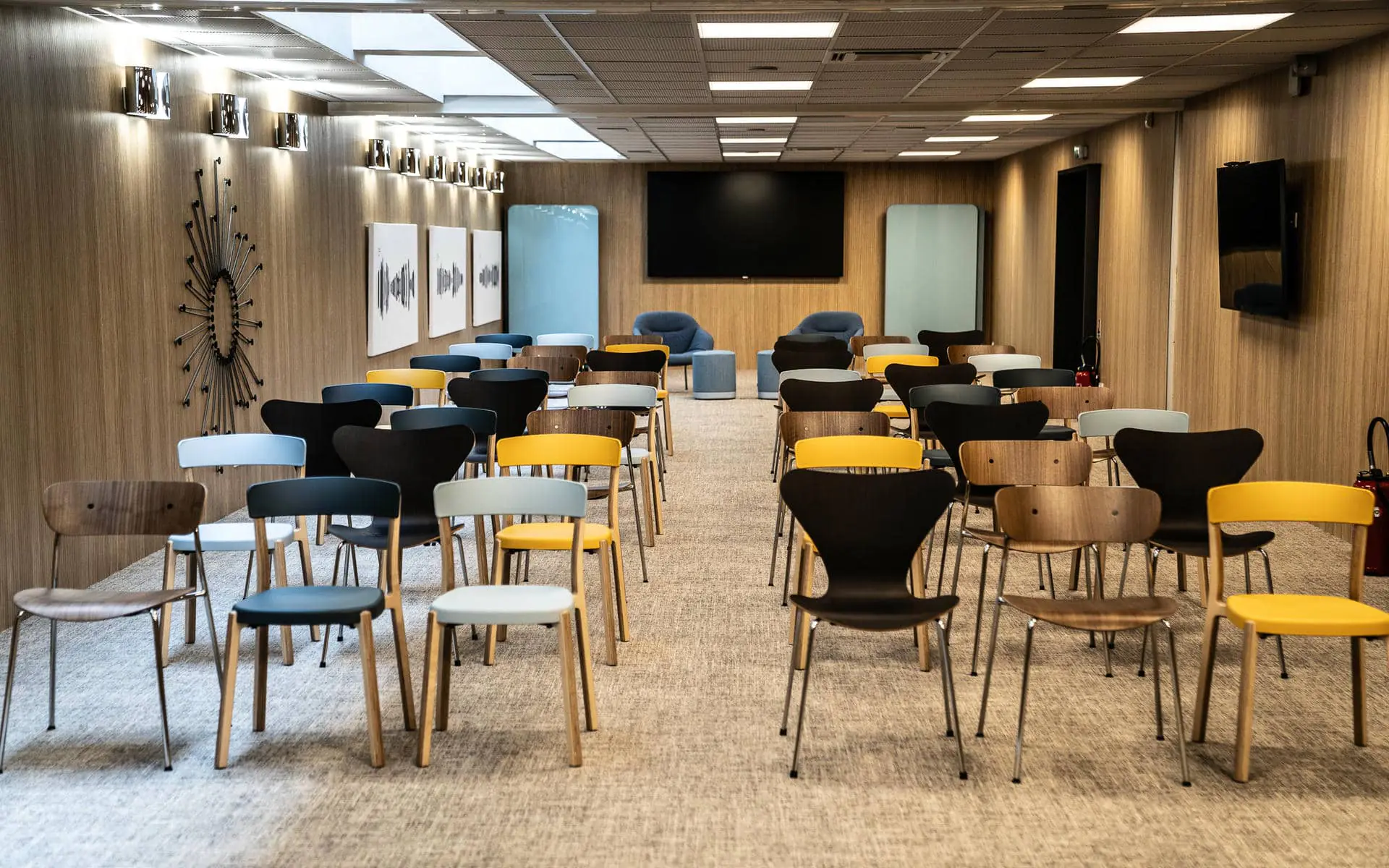 Meeting room rental space with rows of mixed design chairs in yellow, black, grey and wood tones on a beige carpet, wood-panelled walls decorated with a sunburst wall clock and abstract art panels on the left, a large black flat screen at the centre back, two blue armchairs and a frosted glass mobile board in the background, and a suspended tile ceiling with integrated fluorescent lighting.