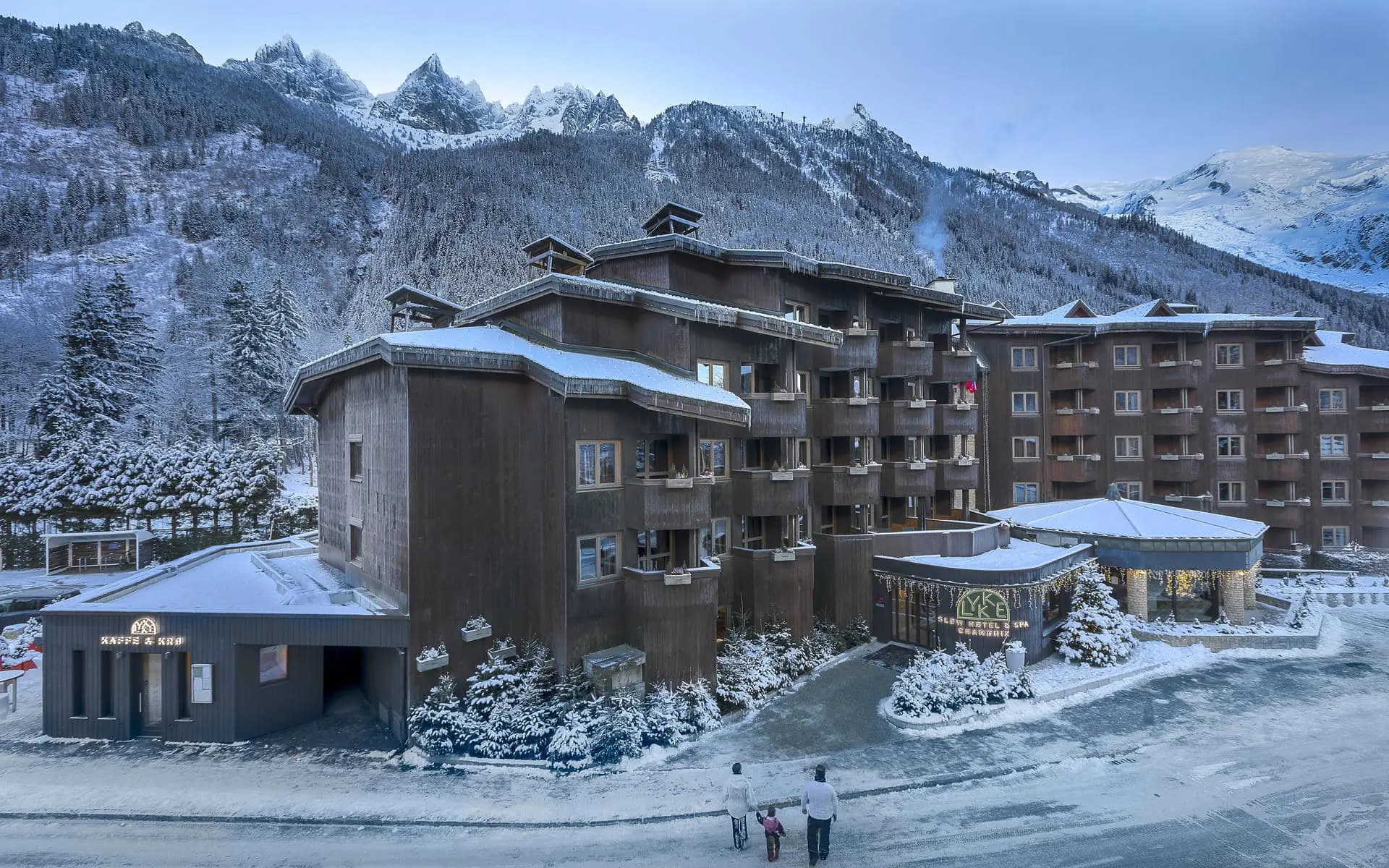 Complexe hôtelier pour un séminaire à la montagne, avec bâtiments en bois dotés de multiples balcons au centre, allées et espaces paysagers enneigés au premier plan, et sommets escarpés enneigés s’étendant sur l’arrière-plan.