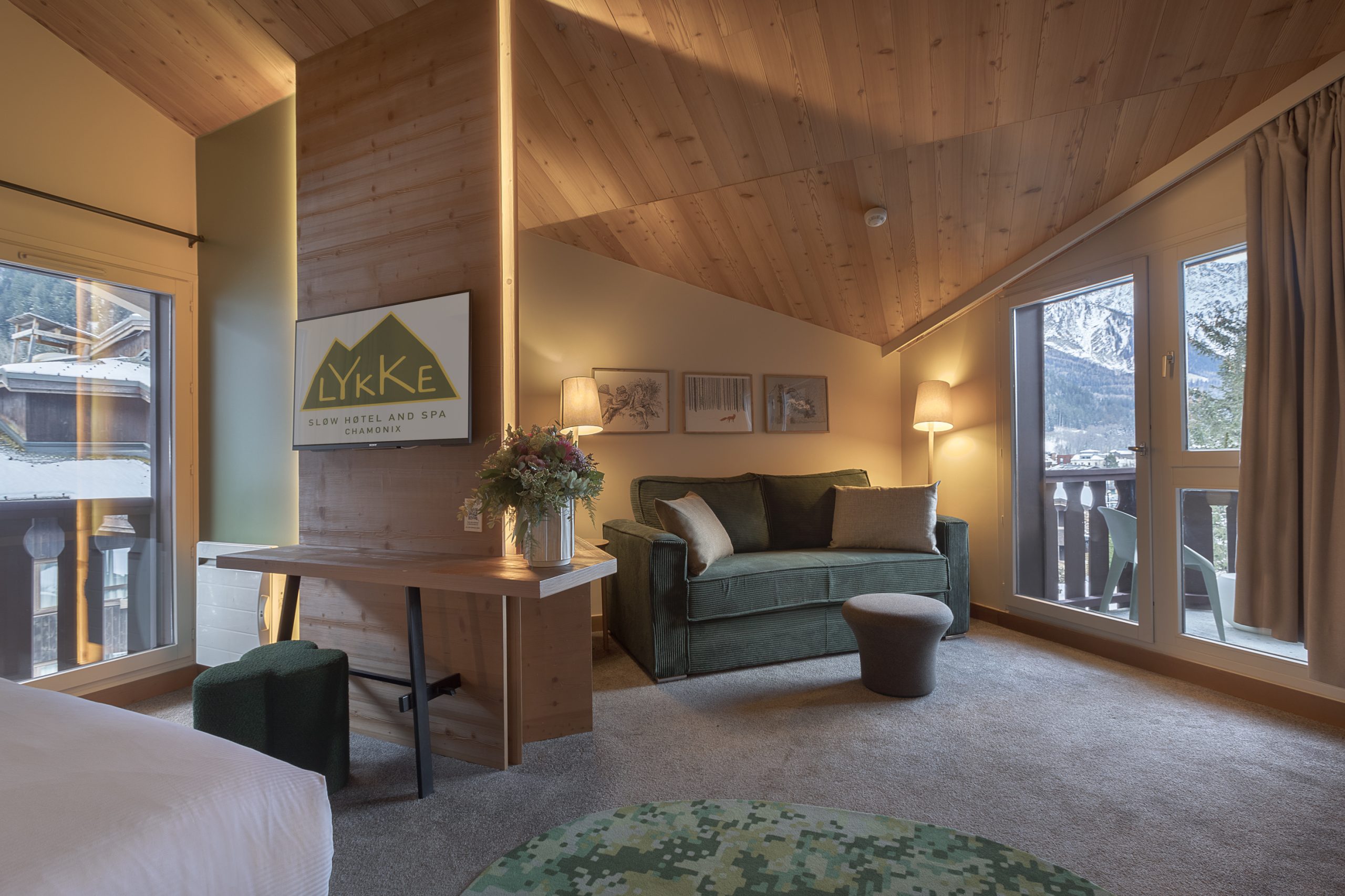 Chambre de l'hôtel Lykke Slow Hotel and Spa avec plafond en pente en bois de pin, comprenant un bureau en bois avec panneau logo de l'hôtel à gauche, un canapé vert matelassé avec bouquet de fleurs et pouf rond au centre, et de larges fenêtres donnant sur un paysage de montagne enneigé en arrière-plan, un cadre inspirant pour un séminaire à la montagne.