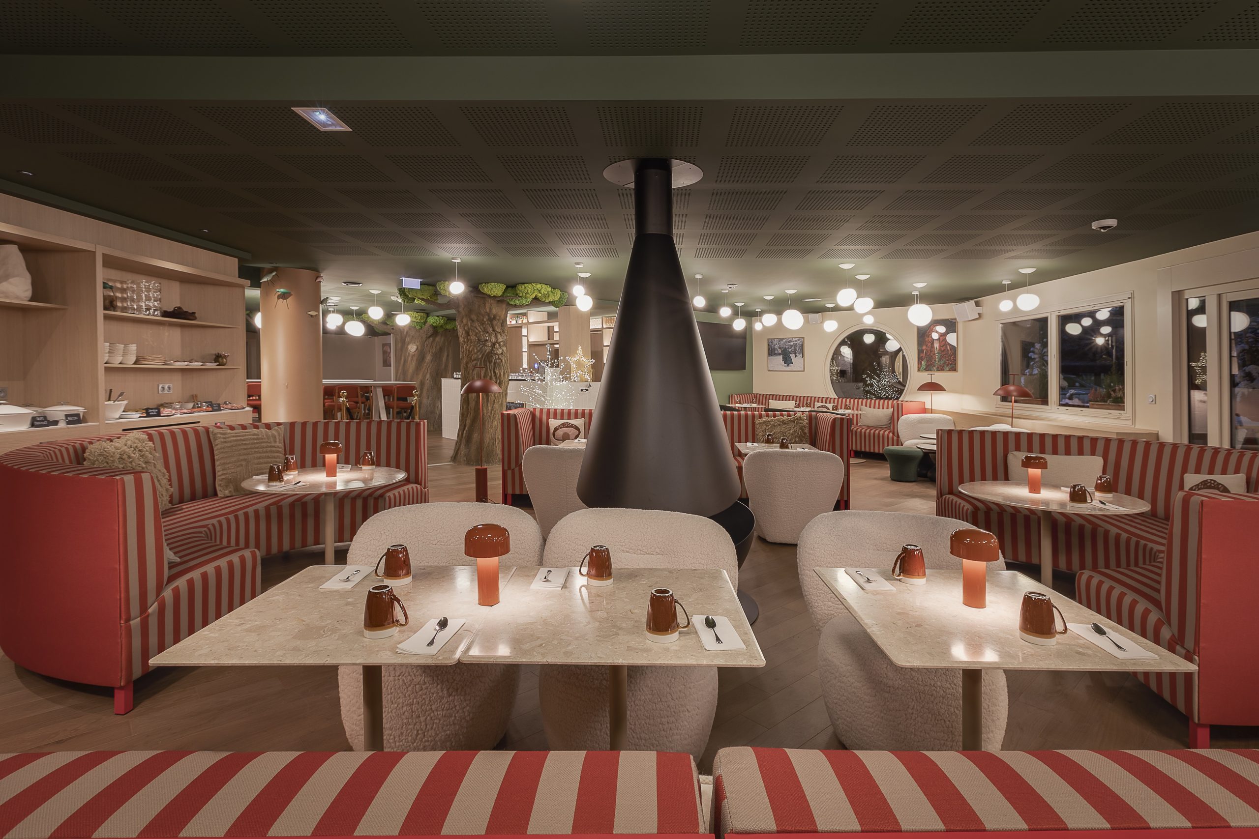 Vue générale d'un restaurant d'hôtel avec plafond vert foncé perforé orné de suspensions globes, comprenant une cheminée suspendue noire en forme de cône au centre, des tables à plateau marbre avec lampes cuivrées et chaises bouclette blanches au premier plan, des banquettes et canapés rayés rouge et blanc dans toute la salle, un tronc d'arbre décoratif en arrière-plan, un hublot circulaire, de grandes fenêtres à droite et un meuble étagère à gauche, pour une restauration sur place.